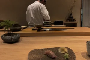 Presents a low-angle, close-up shot from the perspective of a diner seated at a light-colored wooden omakase counter. In the immediate foreground, a single piece of glazed nigiri sushi, likely kinmedai, rests on a textured, dark stone serving plate alongside a small mound of grated wasabi and a pile of pickled ginger. The midground features a thick wooden prep board stretching across the frame, holding several professional Japanese chef knives with dark handles, a small stone bowl containing a delicate green plant, and various ceramic prep dishes. In the softly lit, minimalist background, a sushi chef dressed in a white uniform and traditional white cap stands with his back partially to the camera, meticulously working at his station against a muted, earth-toned wall.
