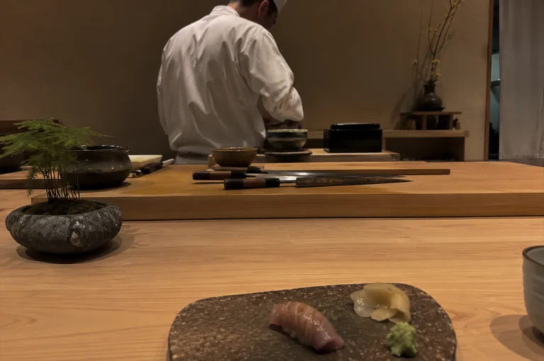 Presents a low-angle, close-up shot from the perspective of a diner seated at a light-colored wooden omakase counter. In the immediate foreground, a single piece of glazed nigiri sushi, likely kinmedai, rests on a textured, dark stone serving plate alongside a small mound of grated wasabi and a pile of pickled ginger. The midground features a thick wooden prep board stretching across the frame, holding several professional Japanese chef knives with dark handles, a small stone bowl containing a delicate green plant, and various ceramic prep dishes. In the softly lit, minimalist background, a sushi chef dressed in a white uniform and traditional white cap stands with his back partially to the camera, meticulously working at his station against a muted, earth-toned wall.