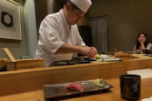 A Japanese sushi chef, wearing a crisp white chef's uniform and a traditional white hat, stands behind a polished light-wood counter meticulously preparing sushi in a dimly lit, minimalist restaurant. In the foreground, a single piece of nigiri topped with a slice of dark red tuna sits on a rectangular, textured ceramic plate alongside small mounds of pickled ginger and wasabi. To the right, a smiling woman sits at the counter with her chin resting on her hand, observing the chef as he works, while a cup of tea sits on the wooden surface in the foreground.