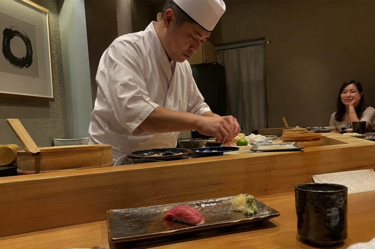 A Japanese sushi chef, wearing a crisp white chef's uniform and a traditional white hat, stands behind a polished light-wood counter meticulously preparing sushi in a dimly lit, minimalist restaurant. In the foreground, a single piece of nigiri topped with a slice of dark red tuna sits on a rectangular, textured ceramic plate alongside small mounds of pickled ginger and wasabi. To the right, a smiling woman sits at the counter with her chin resting on her hand, observing the chef as he works, while a cup of tea sits on the wooden surface in the foreground.