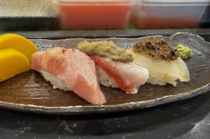 Three pieces of artisanal nigiri sushi are arranged linearly on an elongated, textured dark ceramic plate with a rustic metallic sheen. The left piece features a thick, pale pink cut of fatty tuna (otoro) with delicate marbling; the center piece shows white fish with a visible red stripe, topped with a dollop of greyish-green paste and citrus zest; the right piece displays translucent white fish crowned with a savory, dark minced tapenade. Yellow pickled radish slices sit to the far left, and a small mound of bright green wasabi rests on the far right, set against a blurry background of refrigerated ingredients.