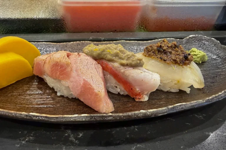 Three pieces of artisanal nigiri sushi are arranged linearly on an elongated, textured dark ceramic plate with a rustic metallic sheen. The left piece features a thick, pale pink cut of fatty tuna (otoro) with delicate marbling; the center piece shows white fish with a visible red stripe, topped with a dollop of greyish-green paste and citrus zest; the right piece displays translucent white fish crowned with a savory, dark minced tapenade. Yellow pickled radish slices sit to the far left, and a small mound of bright green wasabi rests on the far right, set against a blurry background of refrigerated ingredients.