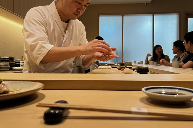 A close-up, low-angle shot from a diner's perspective at an omakase sushi counter features a sushi chef in a crisp white traditional jacket carefully shaping a piece of nigiri with both hands. The light-colored wooden counter in the foreground holds a pair of chopsticks on a ceramic rest and a small dish of soy sauce, while the background shows a minimalist, softly lit dining room where a few customers sit chatting along the extended bar.