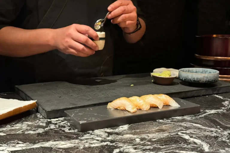 A chef, dressed in a black uniform, is captured mid-action while carefully seasoning a row of four pieces of nigiri sushi laid out on a sleek, dark slate serving board. The background is dimly lit and minimalist, emphasizing the chef's focused hands as they use a small spoon to apply a garnish from a ceramic cup, with a small bowl of wasabi nearby.