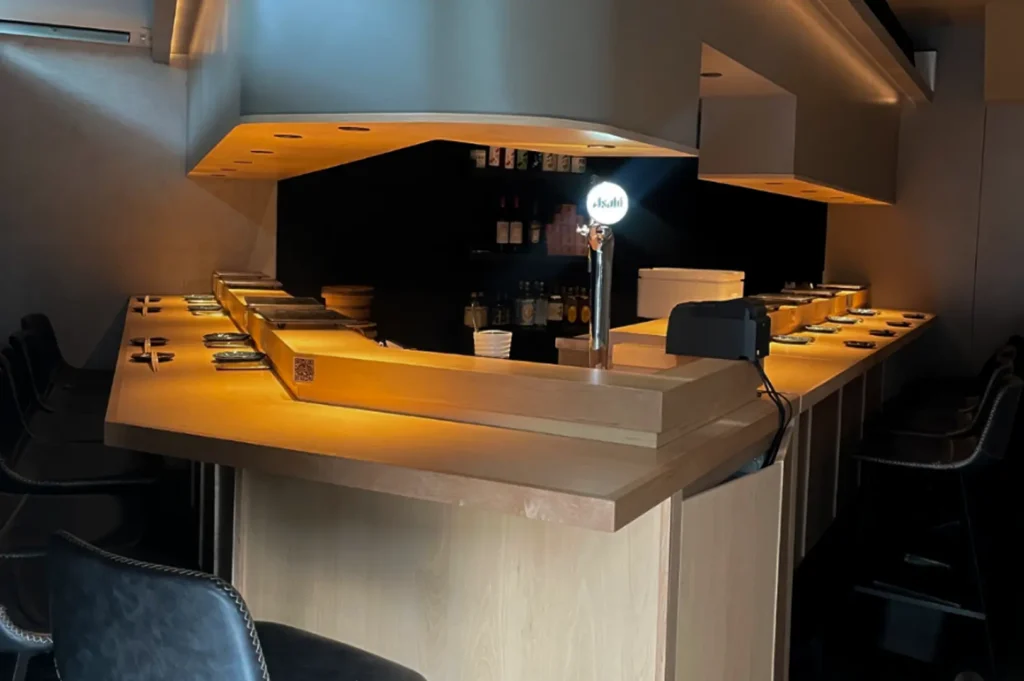 An empty, sophisticated sushi bar is captured in warm, dim lighting, featuring a long, light-wood counter arranged in a U-shape. Each seat is marked by a simple, minimalist place setting, while the background consists of elegant, textured walls and soft, recessed ceiling lights that create a calm, intimate atmosphere perfect for an exclusive dining experience.