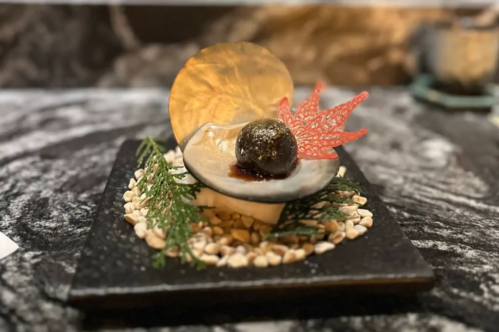 A sophisticated appetizer is meticulously arranged on a square black stone base covered in light-colored pebbles and sprigs of greenery, featuring a single dark, glistening morsel served inside a small shell, topped with a delicate gold disc and a translucent red leaf-shaped garnish.