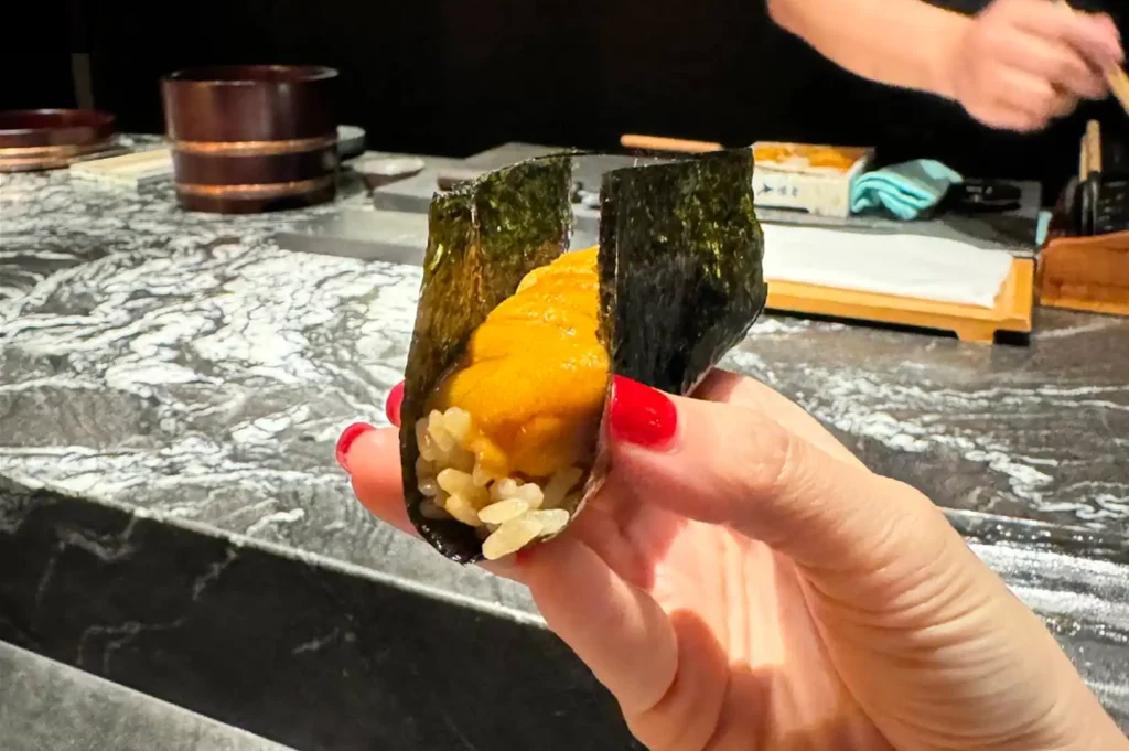 A hand with vibrant red nail polish holds a freshly prepared uni hand roll (temaki), showcasing a generous portion of bright orange sea urchin layered over seasoned rice, all tightly wrapped in a crisp, dark sheet of nori against the backdrop of an omakase counter.