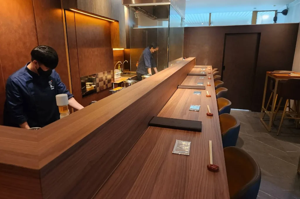 A quiet, minimalist omakase bar is shown in a warm, amber-toned light, featuring a clean, light-wood counter that wraps around the center workspace. An Asahi beer tap stands prominently in the middle of the station, flanked by bottles stored on dark shelves in the back, creating a functional yet aesthetic environment for an intimate dining service.