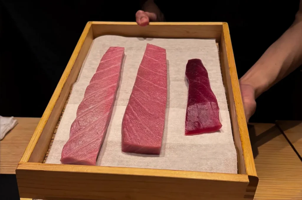 A chef's hands are shown holding a wooden tray lined with white paper, displaying three distinct cuts of raw tuna. The fillets show a color gradient from light pink to a deep, dark red, highlighting the high-quality, fresh, and meticulously prepared fish used in the service.