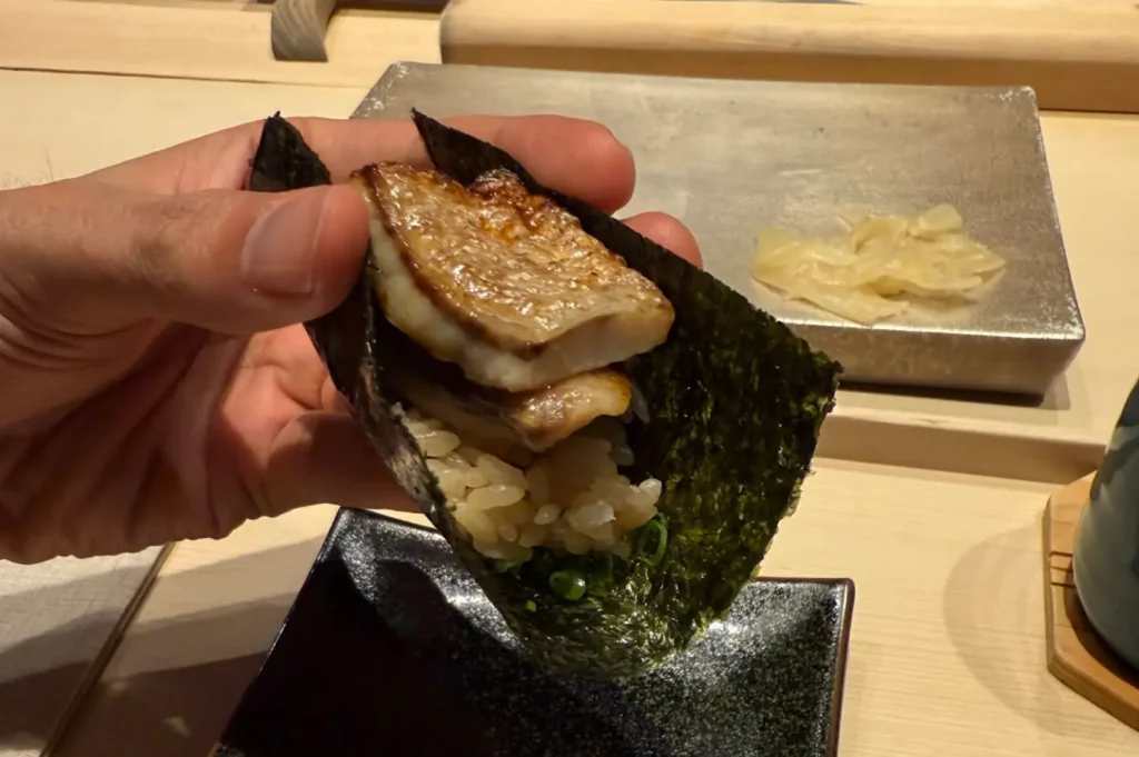 A hand holds a hand-rolled temaki sushi, showcasing layers of seared, golden-brown fish fillets tucked inside a crisp sheet of dark nori. The rice and small bits of green onion are visible at the base of the roll, which is presented against a clean, light-toned wooden table setting.