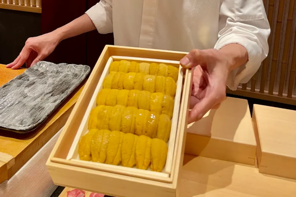 A chef's hands are shown presenting a wooden box filled with rows of fresh, bright yellow uni (sea urchin). The focus is on the uniform, plump texture of the uni, held steady against the warm, minimalist backdrop of an omakase counter.