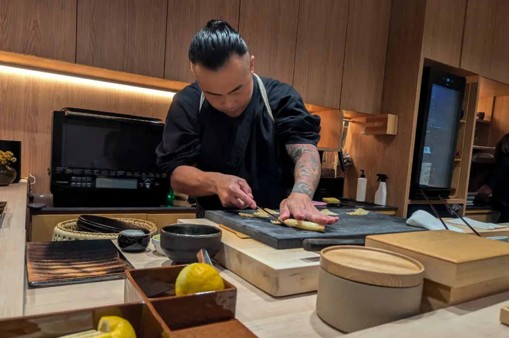 A professional sushi chef with intricate arm tattoos, dressed in a black uniform and apron, is captured in a moment of focused precision as he cuts a piece of fish on a dark wooden board. The setting is a warm, minimalist culinary workspace with sleek, light-wood cabinets, prepared ingredients in ceramic bowls, and soft under-cabinet lighting that highlights the chef's concentrated technique.