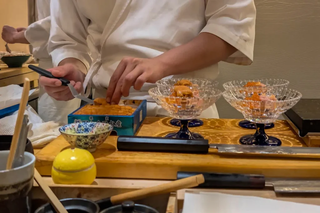 A close-up view at an omakase sushi counter features a chef in a white uniform carefully portioning fresh sea urchin (uni) from a wooden box into small, elegant, clear glass bowls. On the wooden workspace, there are sharp Japanese knives, a small bowl of garnish, and a yellow ceramic container, capturing the meticulous precision involved in preparing high-end sushi.