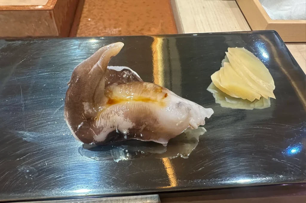 A top-down, close-up shot captures a unique piece of sushi featuring a large, glistening piece of torigai (Japanese heart clam or cockle) on a reflective, dark metallic rectangular serving platter. The clam has a distinct, curved dark brown tail-like protrusion that arches upward and a translucent, fleshy white body brushed with a light, glossy soy glaze down its center. Positioned to the right of the seafood on the same platter is a neat fan of thinly sliced, pale yellow pickled ginger, all resting on a smooth wooden counter surface.