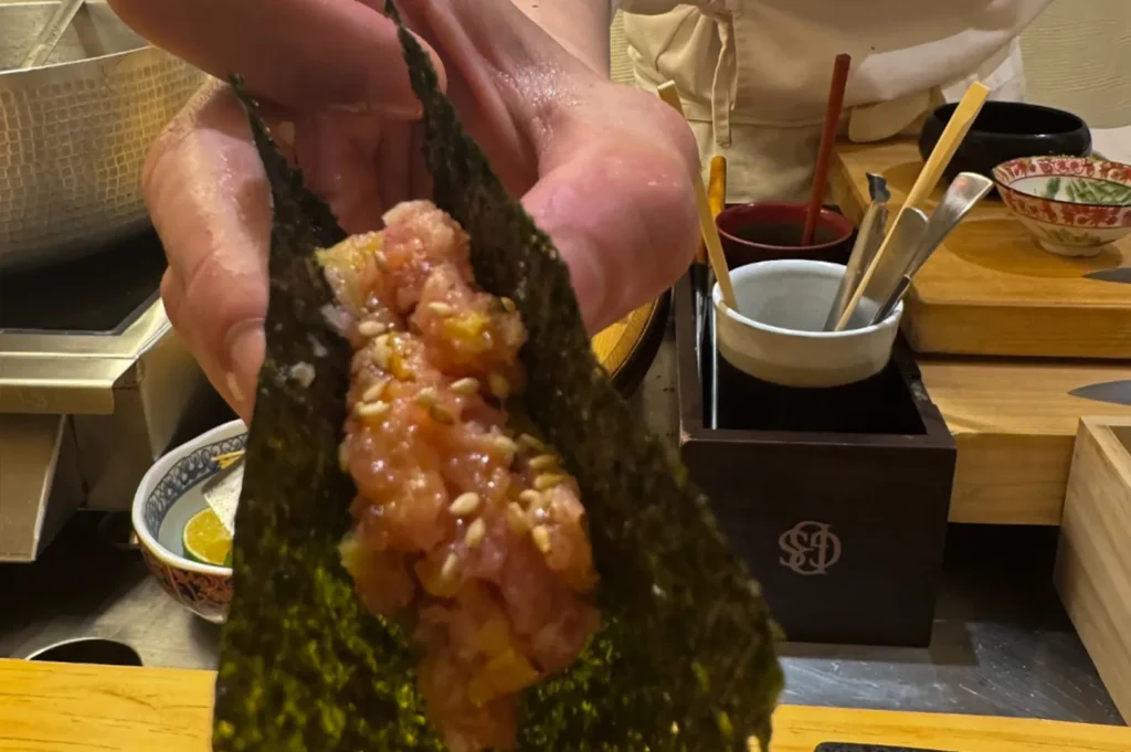 A close-up, first-person perspective captures a chef’s hands holding a piece of hand-rolled sushi, or temaki, wrapped in crispy dark nori. The roll is generously filled with a textured mixture of minced tuna and sesame seeds, highlighting the fresh, handcrafted nature of the preparation as the chef presents it directly to the diner across the wooden counter.