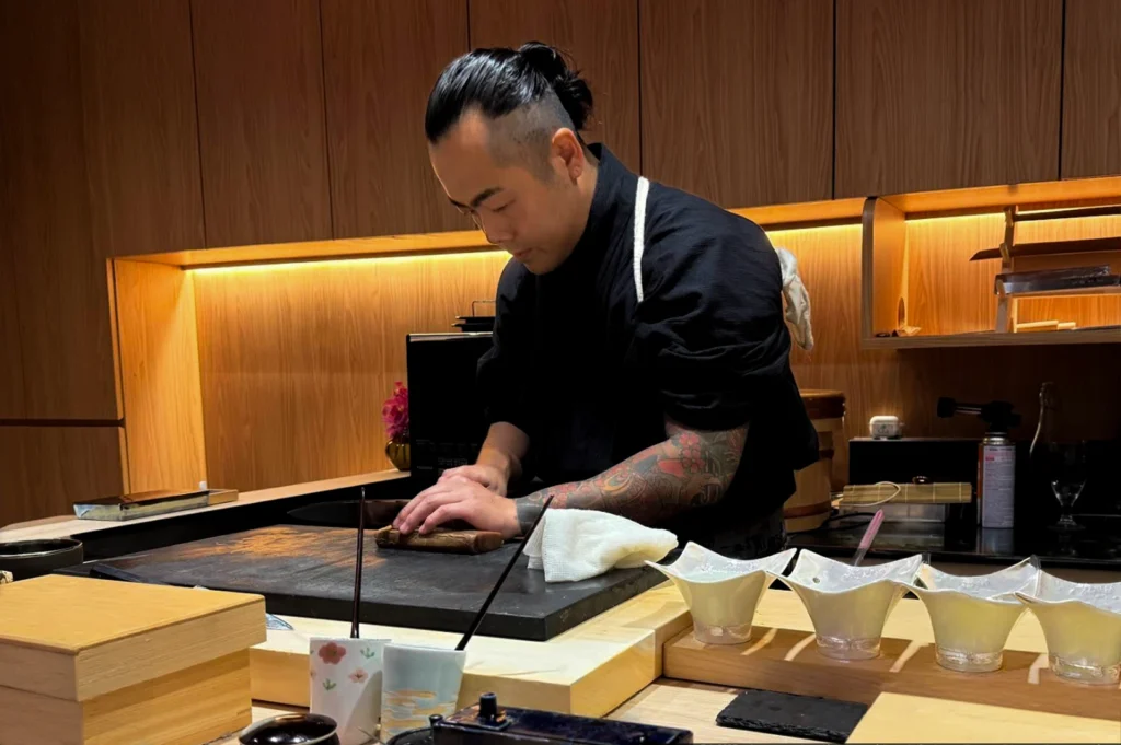 A sushi chef with his hair tied in a bun and visible tattoos on his arms is focused on preparing food behind a wooden counter. He is wearing a black chef’s uniform and is pressing down on a small item with his hands on a dark cutting board. In front of him, several white, star-shaped bowls are arranged on the counter, with wooden surfaces and warm, ambient lighting creating an intimate atmosphere.