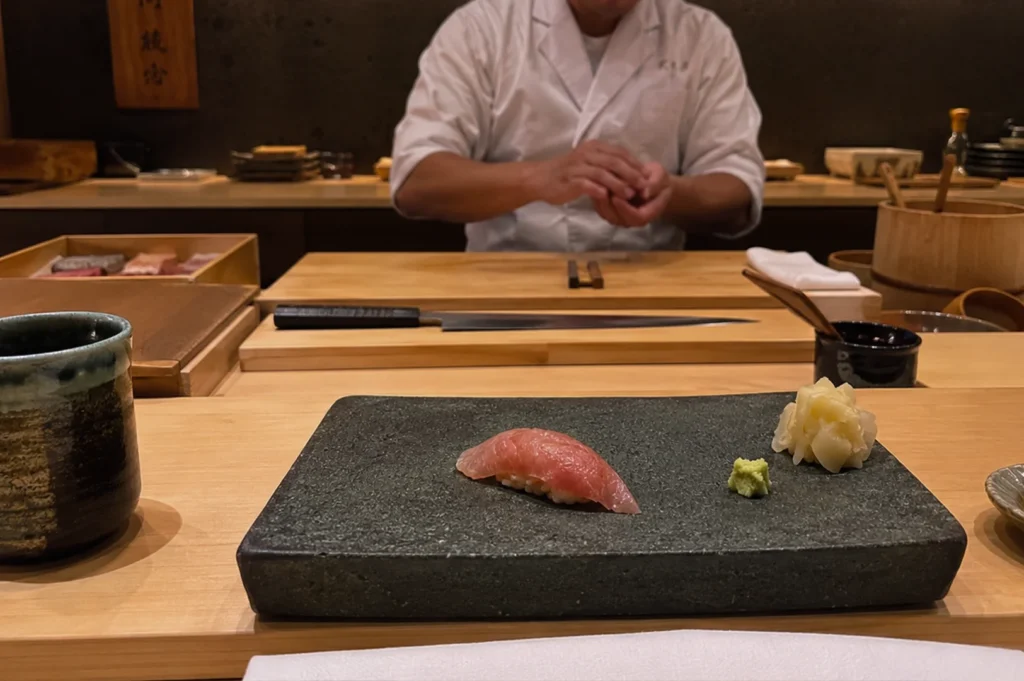 A close-up view of a high-end omakase dining experience features a piece of fresh, pink-hued tuna nigiri served on a dark, rough-hewn stone platter. Next to the nigiri is a small portion of pickled ginger and a dab of vibrant green wasabi. In the background, a sushi chef dressed in a white uniform works behind a pristine light-wood counter, with a long, sharp sushi knife resting on a wooden cutting board, conveying the focused and artisanal atmosphere of the kitchen.