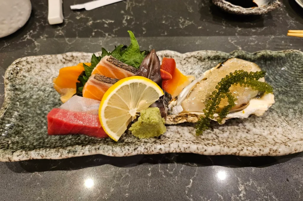 A rectangular, rough-edged ceramic platter with a mottled grey and green glaze presents an elegant assortment of sashimi and fresh seafood. The left side features vibrant cuts of salmon with dark skin, bright red tuna, translucent white fish, and orange surf clam arranged beautifully with green shiso leaves, a fresh lemon wheel, and a dollop of wasabi. The right side of the platter holds a large, opened oyster in its shell, glazed delicately and topped with vibrant strings of green sea grapes (umi-budo), all resting on a dark, speckled stone counter.