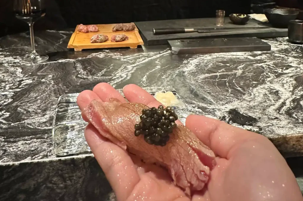 A single piece of premium nigiri sushi, topped with a generous dollop of black caviar, is held delicately between two hands. The background reveals a blurred, luxurious dining setting featuring a dark, swirling marble countertop, a wine glass, and a small wooden board holding other small portions of food, highlighting the intimate and hands-on nature of the omakase service.