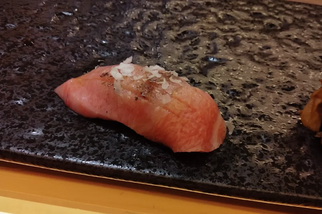 A single piece of premium fatty tuna sushi rests on a textured, dark, obsidian-like slate serving plate. The nigiri is topped with a delicate garnish of sea salt and perhaps a hint of citrus or shiso, highlighting the minimalist and artistic presentation typical of a refined omakase dining experience.