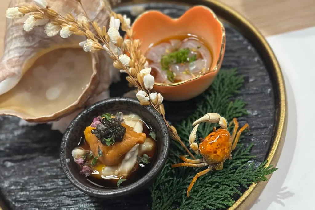 An overhead close-up showcases an exquisite seafood assortment meticulously arranged on a round, textured black platter with a gold rim. The presentation features a small black bowl filled with creamy orange sea urchin (uni) topped with black caviar and tiny purple flowers, alongside a small orange, heart-shaped bowl containing delicate, translucent ribbons of white fish garnished with chopped chives. A large, spiral seashell rests on the left side, while a whole, small roasted crab is positioned on the right over a bed of green cedar sprigs, all tied together by a decorative stalk of dried rice grains arching across the center.