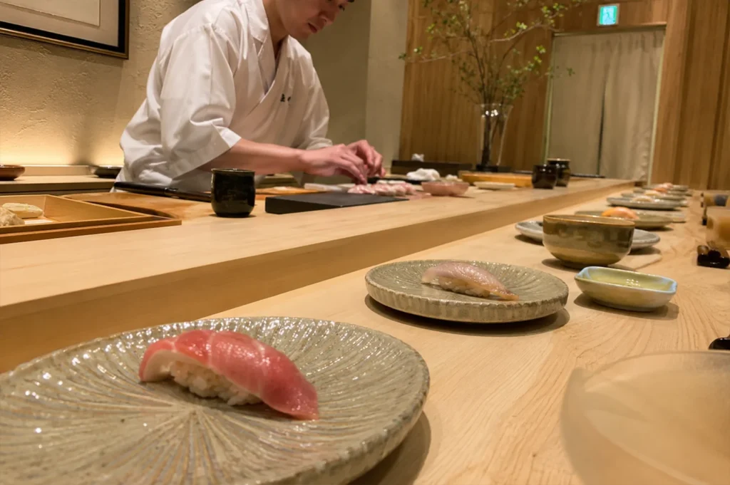 A low-angle, wide perspective looking down a long, light-colored wooden omakase counter highlights a row of identical, textured ceramic plates stretching into the distance, each holding a fresh piece of nigiri sushi. In the immediate foreground, a plate presents a vibrant pink piece of tuna nigiri, while the subsequent plates hold lighter, translucent fish varieties. In the background, a sushi chef in a crisp white uniform is meticulously preparing food, framed by minimalist Japanese decor that includes light wooden wall panels, a tall glass vase with delicate green branches, and an illuminated exit sign over a doorway.