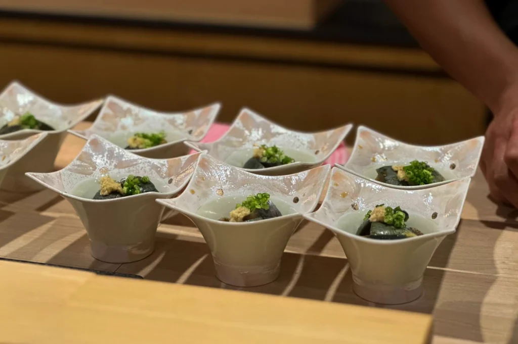 Several small, elegant, star-shaped ceramic bowls are lined up on a wooden counter, each containing a dark ingredient topped with a garnish of ginger and finely chopped green onions. The bowls feature a delicate, light-colored speckled design, and the focus is sharp on the foreground, while a person’s hand is partially visible on the right side of the frame.