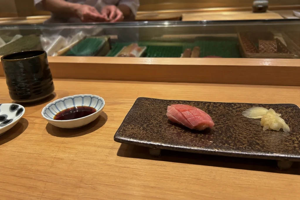 A single piece of tuna nigiri, featuring intricate knife work on the fish, is presented elegantly on a long, dark, earth-toned ceramic plate. Beside the sushi, there is a small mound of pickled ginger, and to the left of the plate, a small decorative bowl contains soy sauce. In the blurred background, a chef in a white uniform is visible behind a wooden counter, highlighting the intimate and prepared nature of an omakase meal.