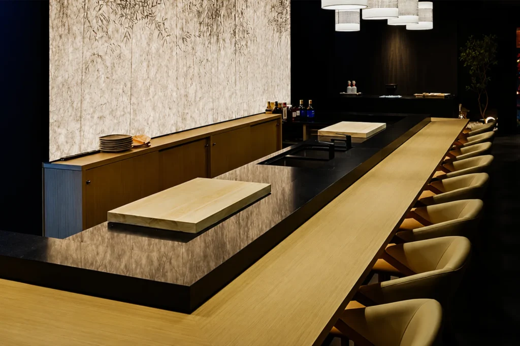 An empty, upscale omakase sushi bar is viewed from a high angle, showcasing a sleek, L-shaped wooden dining counter lined with minimalist, cream-colored leather upholstered chairs. Behind the counter sits an elevated black prep area containing inset sinks and two large, light-wood cutting boards positioned for chefs. The back wall features a striking, softly illuminated panel textured with a subtle black-and-white bamboo forest motif, complemented by modern, cylindrical white woven pendant lights hanging from the ceiling to create a serene and luxurious ambiance.