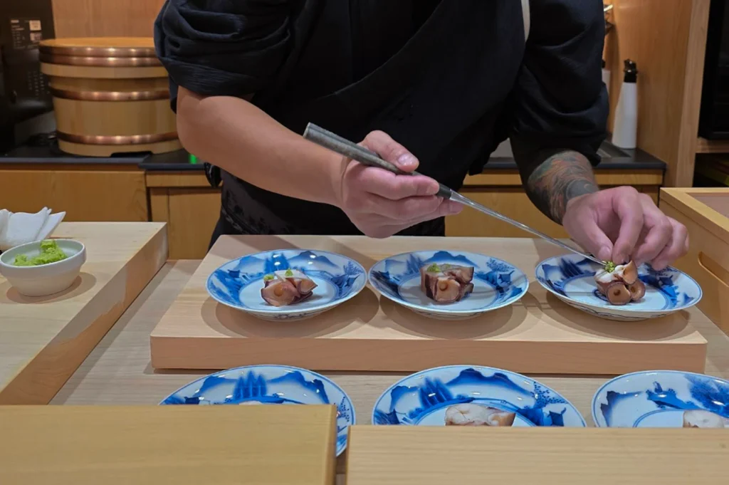 A close-up shot focuses on a sushi chef’s hands and forearms, revealing a detailed colorful tattoo sleeve, as he uses long, slender metal plating chopsticks to delicately place a small garnish onto sliced octopus tentacles. Three elegant blue-and-white patterned ceramic dishes are arranged on a light wood prep block, each holding a meticulously prepared portion of octopus topped with a touch of wasabi.