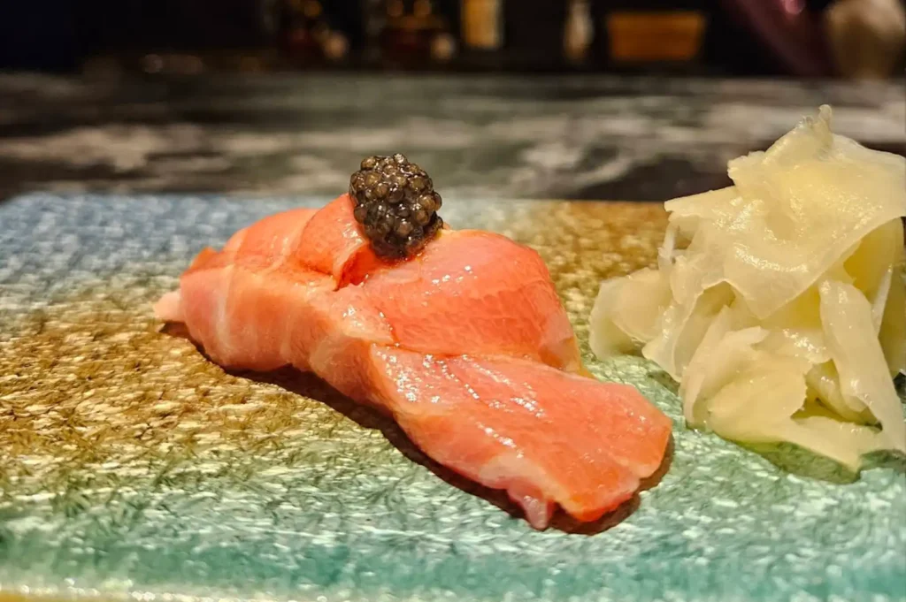 A close-up view of a high-quality piece of fatty tuna nigiri sits on a textured, light-blue plate next to a small, neat mound of pickled ginger. The fish has a rich, marbled pink appearance and is elegantly topped with a small, glistening scoop of black caviar, emphasizing the precision and premium ingredients characteristic of an omakase experience.
