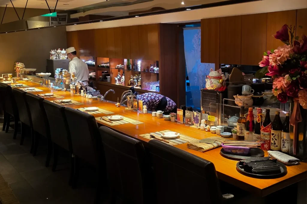 This shot of an elegant omakase bar captures a long, curved counter made of light-colored wood, stretching across the frame to create a warm, inviting dining space. The counter is meticulously set with individual place settings and chopsticks, and the background features shelves displaying bottles of sake and Japanese ceramics, all illuminated by soft, focused lighting that creates a sophisticated ambiance.