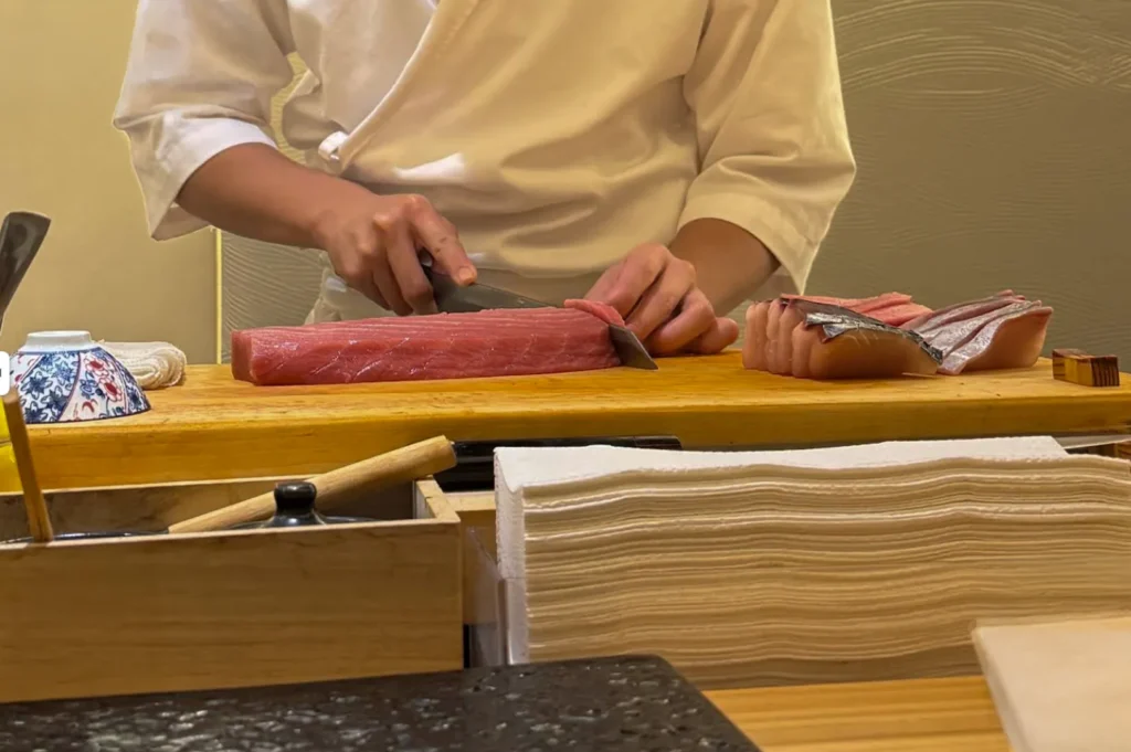 A chef dressed in a crisp white uniform works behind a long, light-wood sushi counter, focusing intently on slicing a large, rectangular block of deep-red tuna. To the right, various portions of prepared sashimi are neatly arranged on the wooden board, showcasing the skilled technique and preparation required for traditional omakase service.