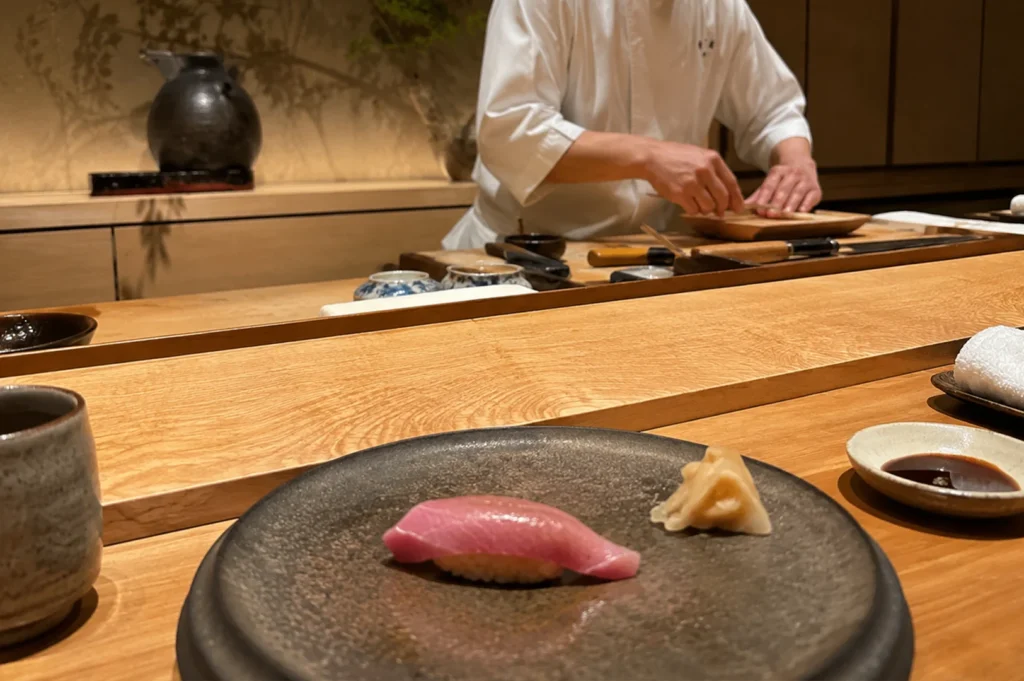 A close-up view from a diner's seat showcases a single piece of glossy, medium-pink tuna nigiri sushi arranged on a round, textured dark grey ceramic plate with a small pile of pickled ginger to its right. A small dish of soy sauce and a stone tea cup flank the plate on a wide, beautifully grained wooden counter. In the softly focused background, a sushi chef in a white uniform operates behind the elevated prep station, which holds traditional wooden blocks and long sushi knives, set against an elegant gold-toned wall featuring a faint, artistic tree branch mural and a large ceramic vessel.