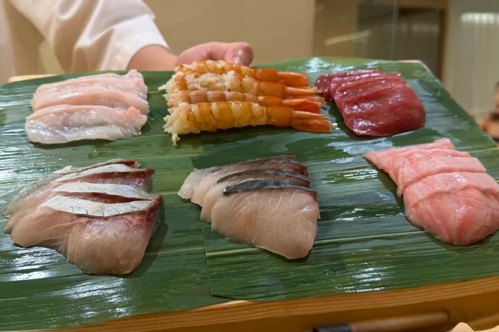 A professional omakase chef, wearing a clean white uniform and a black face mask, stands behind a light-wood counter preparing a meal. In front of them are various fresh seafood ingredients on a wooden board, a chef's knife, a black torch for searing, and a menu placed near a tall glass of iced drink, capturing the focused and methodical environment of the restaurant's kitchen station.