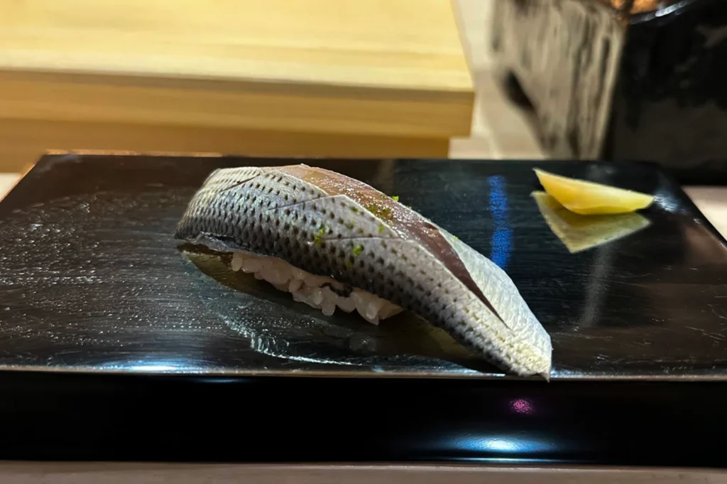 A single piece of nigiri sushi is presented on a rectangular black plate. It features a piece of silvery, skin-on fish draped over a small mound of vinegared rice, with a light dusting of green seasoning visible on the surface of the fish. A small sliver of yellow pickled ginger sits to the right of the sushi on the polished, reflective black surface.
