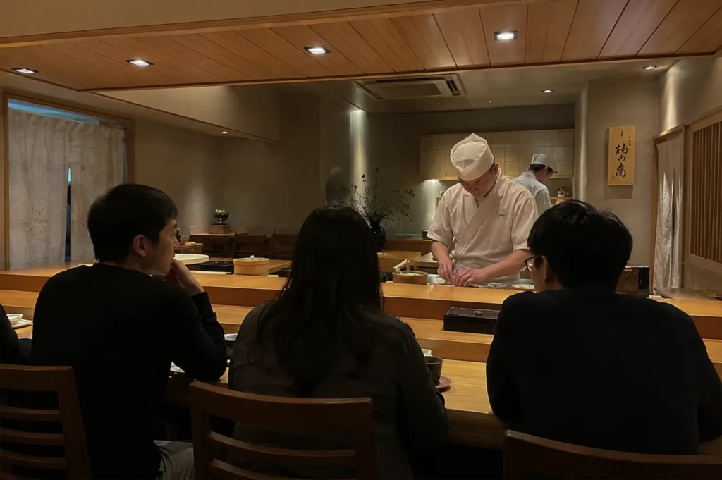 Three patrons are seated at a wooden counter in a quiet, warmly lit Japanese restaurant, viewed from behind as they watch a sushi chef in a white uniform prepare their next course. The chef stands behind the clean wooden prep station, focused on his craft, while the diners wait in anticipation. The scene emphasizes the direct, personal connection between the chef and the guests that is hallmark to the omakase dining style.