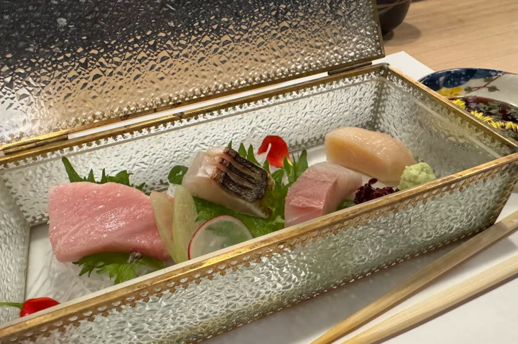 An exquisite selection of sashimi is presented inside an open, rectangular glass box featuring a vintage, gold-embossed filigree trim along its edges and a textured, ice-like finish on its panels. Inside, resting on a bed of shredded white radish and green shiso leaves, are premium pieces of raw fish, including fatty pink tuna, seared mackerel, pale white fish, and a plump scallop accompanied by a tiny red flower petal and a dollop of wasabi. Wooden chopsticks rest on the white tabletop in front of the box, with a small blue ceramic soy sauce dish partially visible in the upper right.