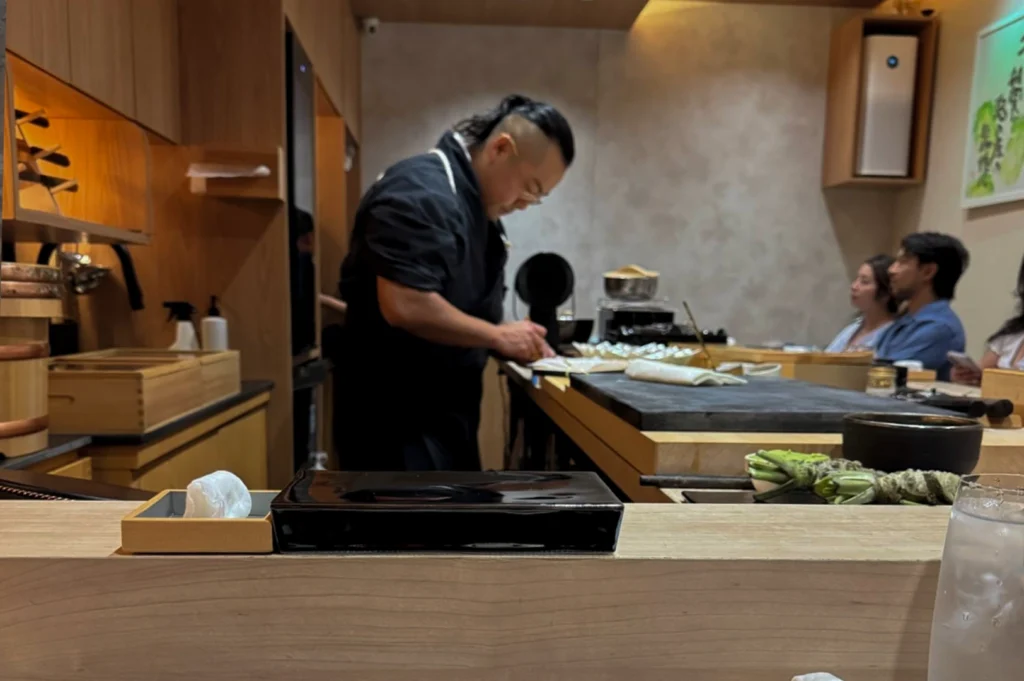 A wide-angle view of a modern, minimalist omakase restaurant reveals a chef with a distinctive shaved-side hairstyle and a black uniform preparing food behind a wide wooden counter. In the foreground, a sleek black rectangular serving plate and a small wooden box with a hand towel await the diner, while several fresh real wasabi roots rest on the counter next to a bowl, and customers are seen enjoying their evening in the dimly lit background.