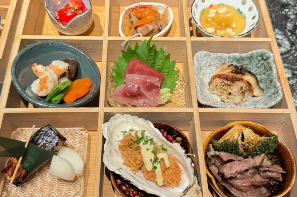 A beautiful wooden bento-style box is divided into nine compartments, each holding a different, meticulously prepared Japanese appetizer or dish. The variety includes vibrant items like grilled seafood, fresh sashimi served on a shiso leaf, cooked vegetables, a crispy fried dish topped with a creamy sauce, and thinly sliced grilled meat, showcasing the diverse and aesthetic presentation of a multi-course meal.