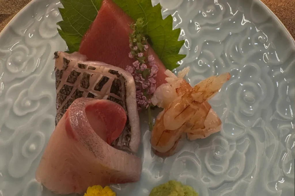 A beautiful plate of sashimi is presented on a light blue, textured ceramic dish, featuring an assortment of thinly sliced raw fish, including tuna and white fish. The arrangement is garnished with a fresh green shiso leaf, small purple blossoms, a dab of bright green wasabi, and yellow edible flowers, emphasizing the visual artistry of the course.