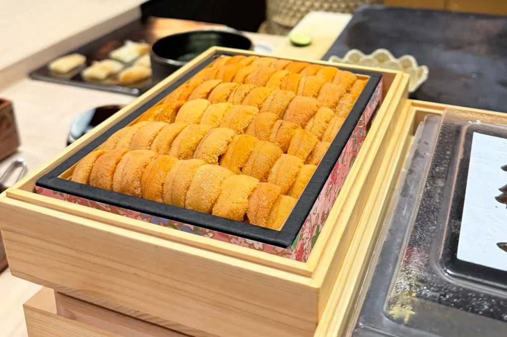 A close-up, angled shot highlights a traditional wooden box filled to the brim with rows of premium, bright orange sea urchin (uni). The pieces of uni are meticulously laid out in overlapping, uniform columns, showcasing their delicate, bumpy texture and vibrant color. The inner rim of the box is lined with a decorative pink floral border, and the container is tilted slightly forward on a light-colored wooden sushi bar, with obscured plates and kitchen elements softly blurred in the background.