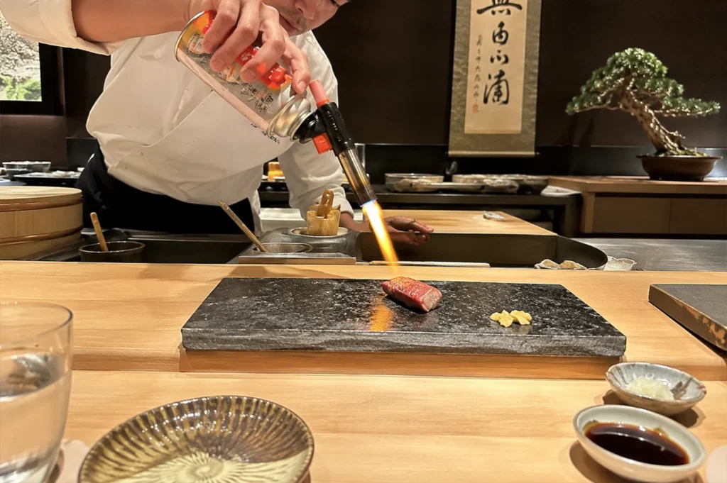 A chef in a white uniform uses a handheld blowtorch to sear a single piece of marbled wagyu beef or fatty tuna sushi, creating a bright orange flame that reflects intensely off the smooth, dark stone slab beneath it. The stone platter also holds a small pile of grated ginger or garlic, sitting on a light-colored wooden counter alongside a striped ceramic plate, a dish of soy sauce, a small bowl of green garnishes, and a water glass. The background features authentic Japanese decor, including a hanging scroll with black calligraphy, a dark wooden shelf, and a carefully manicured bonsai tree in a round pot.