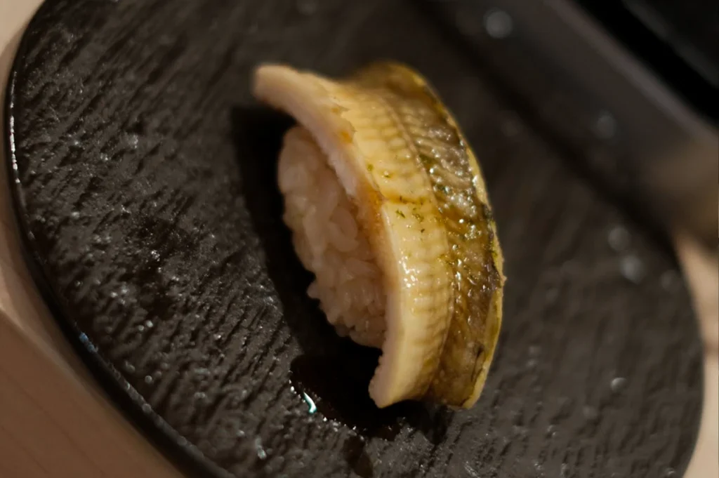 A piece of nigiri sushi rests on a textured black plate, featuring a slice of cooked, light-colored fish with visible skin texture layered over a mound of rice. The fish appears glazed with a subtle sheen, and a small drop of dark sauce is visible on the plate's dark, grooved surface just below the sushi.