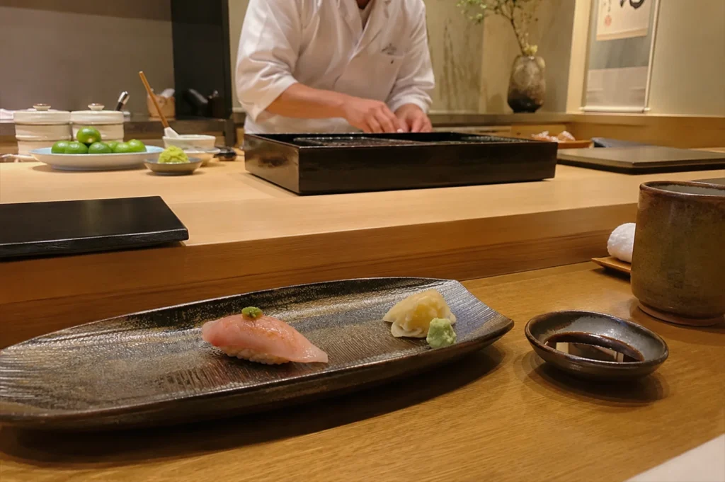 A piece of pale pink nigiri topped with a tiny spot of wasabi rests on a long, textured, dark ceramic plate in the foreground of a sushi bar. Beside it are small portions of pickled ginger and wasabi, and a small dish of soy sauce. The background features a blurred, out-of-focus view of a sushi chef in a white uniform working behind a light-wood counter, with a plate of small citrus fruits and other garnishes neatly arranged on the surface.