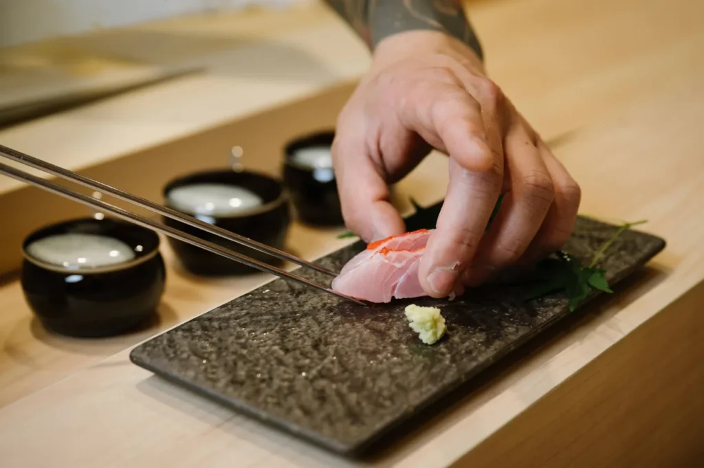 A close-up shot captures the hand of a sushi chef delicately placing a piece of white-fish nigiri sushi onto a textured, dark grey rectangular plate. Long, slender metal plating tweezers are held in the chef's other hand, assisting in the precise arrangement next to a small mound of fresh wasabi. In the softly blurred background on the light wooden counter sit three small, glossy black ceramic bowls filled with clear liquids, emphasizing the focus on the chef's culinary craftsmanship.