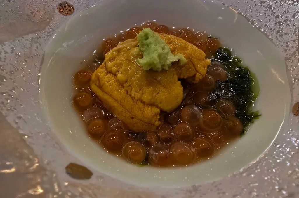 A top-down macro shot showcases an exquisite culinary dish presented in a textured, clear glass bowl with gold flecks. The dish features a generous, vibrant orange piece of uni (sea urchin) topped with a dollop of bright green fresh wasabi, resting on a bed of glistening, translucent ikura (salmon roe) marinated in a delicate, dark broth with a side of sea vegetables.