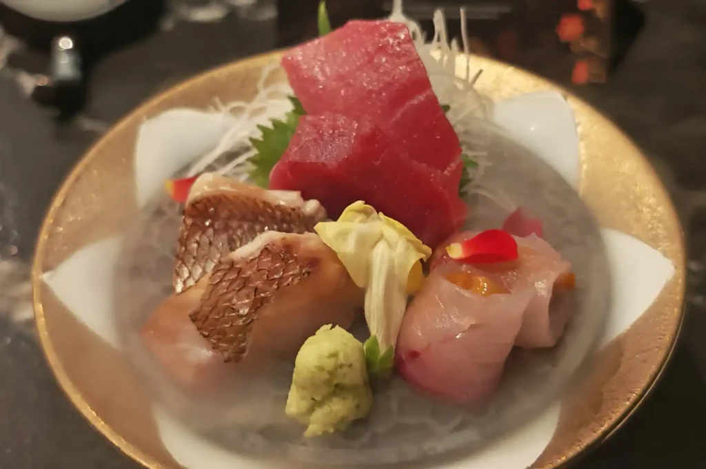A platter of assorted sashimi is presented in a shallow, gold-rimmed bowl surrounded by a layer of misty dry ice, which creates an ethereal, smoky effect. The selection includes a large, deep red piece of tuna, seared fish with visible skin texture, and a delicate roll of white fish topped with a hint of orange roe, all garnished with fresh wasabi and edible flower petals.
