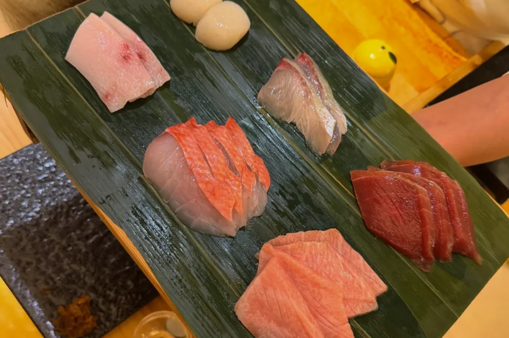 An overhead, slightly angled shot shows a dark green bamboo leaf holding a variety of fresh sashimi slices served on a wooden platter. The selection includes different cuts of tuna, white fish, and plump scallops, all laid out with care, illustrating the diverse and high-quality ingredients featured in an omakase meal.