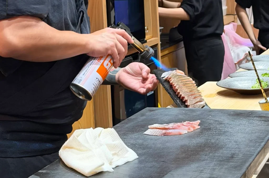 A close-up shot focuses on the hands of a chef wearing a dark gray uniform as he uses an orange-and-white canister blowtorch to lightly sear food. With his left hand, he aims a bright blue flame at a long line of sliced fish arranged on a dark slate platter held aloft, while several pieces of raw, pale pink fish with distinct white scales wait on a large dark cutting board in the foreground. A crumpled white towel rests on the corner of the workspace, and the background shows a bustling restaurant kitchen with other staff members dressed in black uniforms moving past light wooden cabinetry.